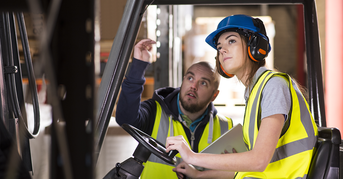 A young white woman is learning how to use a forklift and she is being instructed by a colleague who has a binder in hand. Both are wearing bright neon yellow safety vests and the young woman is also wearing sound-muffling headphones.
