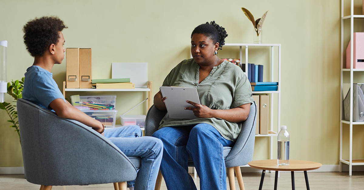 A Black teenage boy sits across from a Black female counselor.