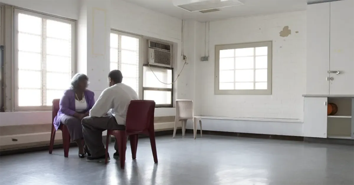 A woman ad a man sit in a sterile, otherwise empty room in a prison, talking across a table.