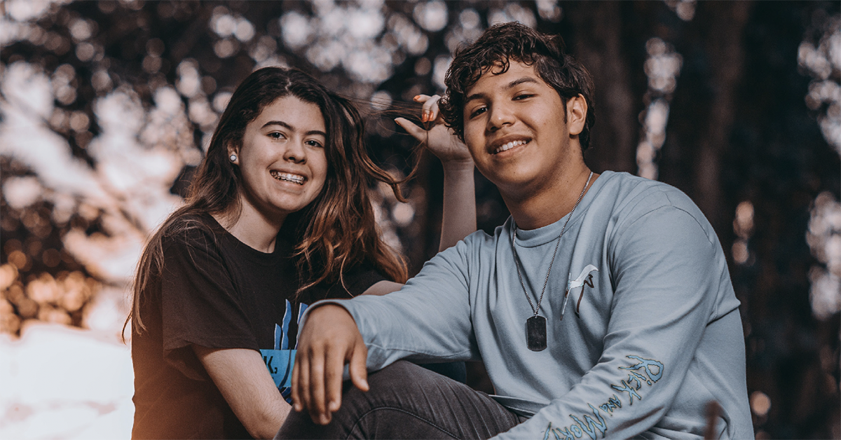 Two young teenagers look at the camera and smile. They are seated outdoors with a sunset or sunrise and the shadow of trees behind them.