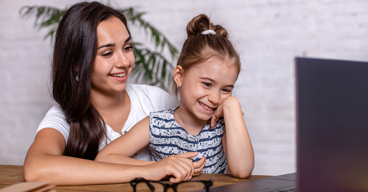 A young mother sits with her daughter on her lap. They are in an office setting, looking at a laptop, and smiling at the same time.