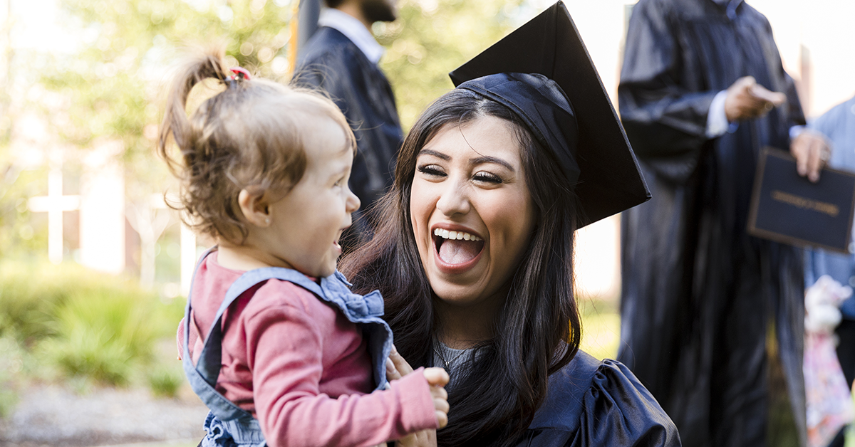 A young woman smiles while wearing a graduation cap and gown and she's holding her toddler daugther.