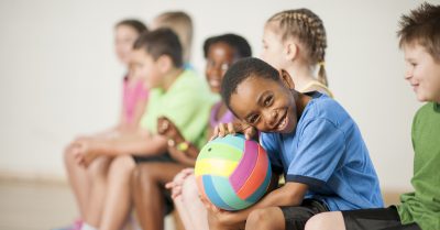 A primary school boy sits at gym class with a colorful volleyball in his lap. He smiles at the camera with his classmates sitting in a line around him.
