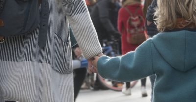 A woman holds a young child's hand as they walk through a crowd. Both mother and child have their back to the camera.
