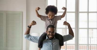 A young Black father stands with his young son sitting atop his shoulders. The two are smiling with their arms in the air.