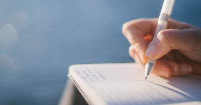 A hand holding a pen sits above an open notebook.