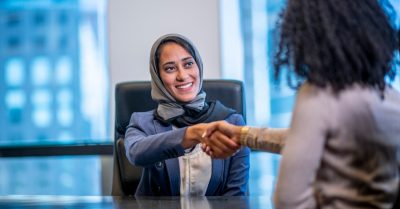 Picture of woman, wearing a headscarf and blazer, shaking the hand of another woman in an office setting.