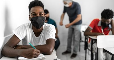 A young black man sits at a desk in a classroom. He's wearing a mask.