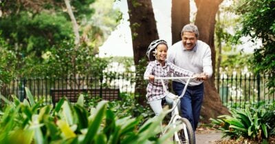 An Afro-Latino grandfather helps his grandaughter ride a bike through a lush springtime park. They smile as she pedals and he steadies the bike's handlebars.