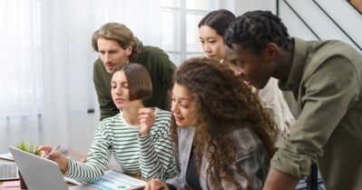 A group of five young people of various races and ethnicities look at a laptop on a table.