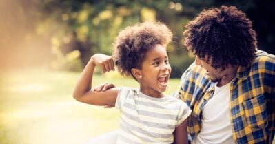 A young schoolgirl sits next to her parent, smiling wide, as she flexes a bicep to show how strong she is.