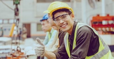 In a factory setting, a young Asian man smiles and gives a thumbs-up sign while wearing a hard hat, gloves and a reflective vest.