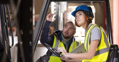 A young white woman is learning how to use a forklift and she is being instructed by a colleague who has a binder in hand. Both are wearing bright neon yellow safety vests and the young woman is also wearing sound-muffling headphones.