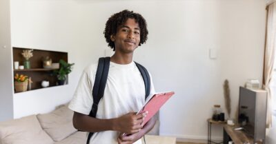 A young person wearing a backpack holds a pink notebook and smiles at the camera.