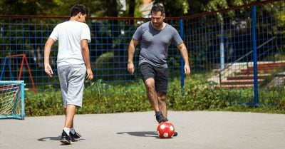 And older man and a older boy on pavement, playing soccer.