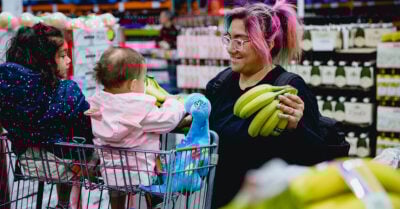 A parent with two young children shops for groceries, smiling as they hold bunches of bananas at a supermarket.