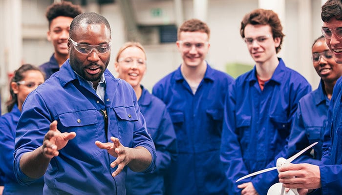 A teacher instructs his class of young adults in an engineering workshop. They are all wearing protective eyewear and blue coveralls.