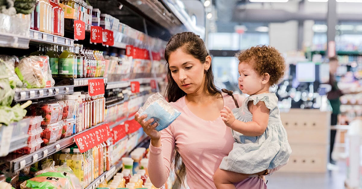A woman holding a young child shops in a grocery store, examining a food item while standing in an aisle lined with products and sale signs.