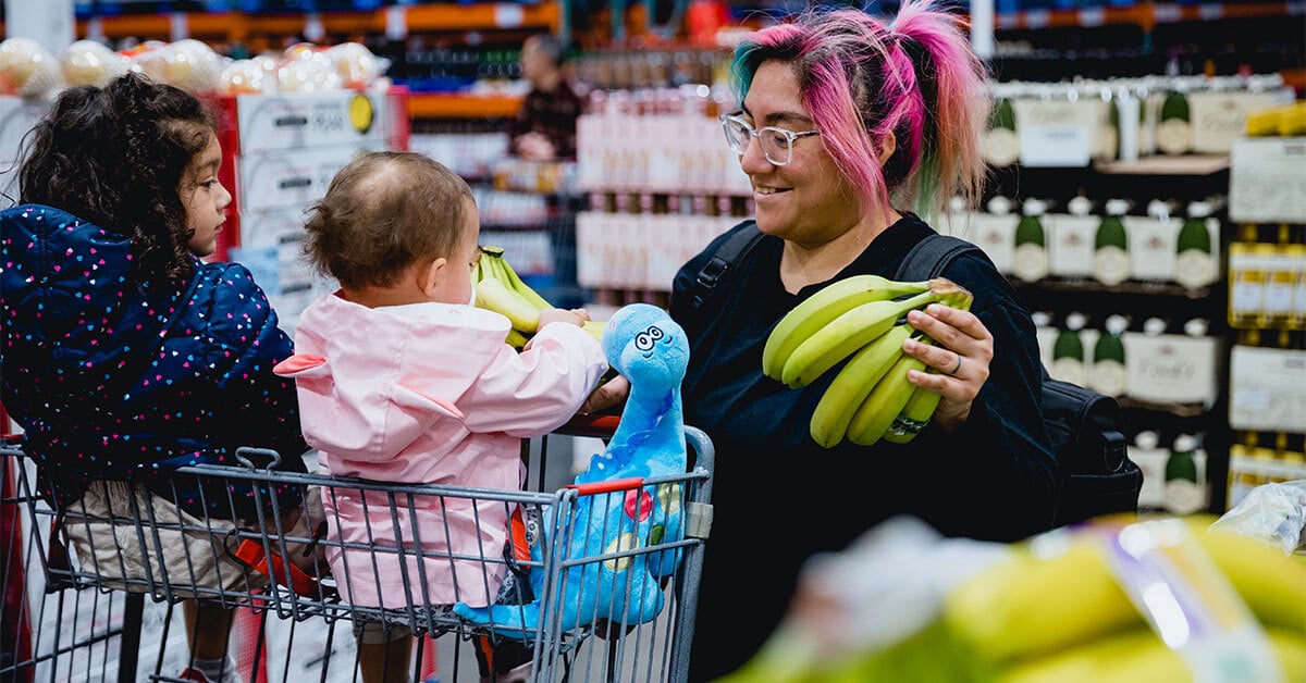 A parent with two young children shops for groceries, smiling as they hold bunches of bananas at a supermarket.