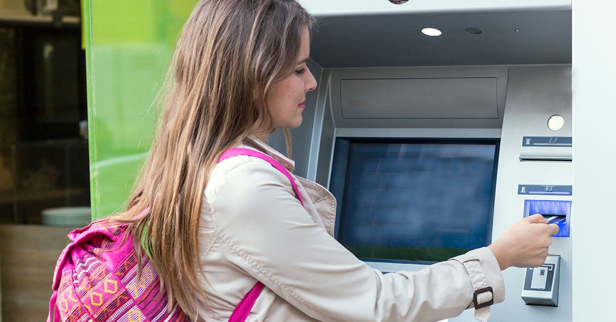 Teenage girl with blonde hair, coat and backpack is using an ATM machine.