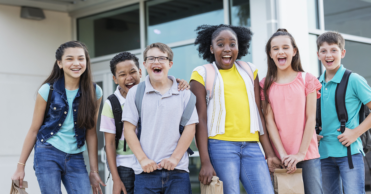 A diverse group of elementary-aged kids wearing backpacks stand outside a building smiling