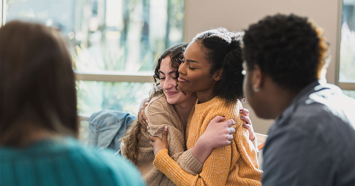 Two young people share a comforting embrace during a group conversation, while others sit nearby offering support.