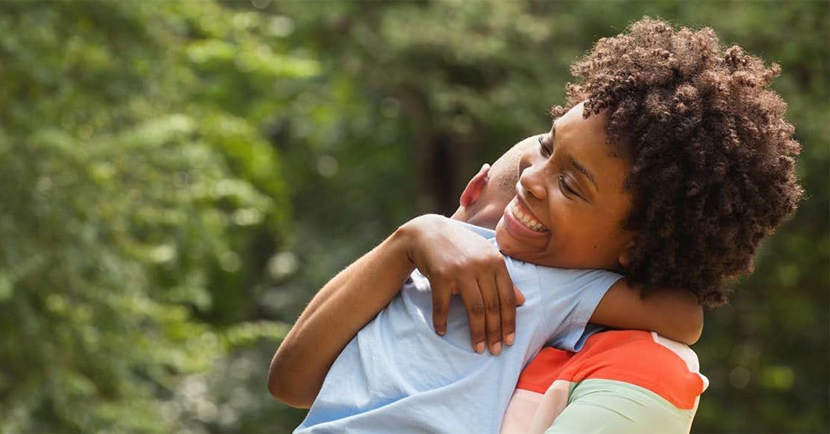 Black mother smiling and standing outside as she holds her young son to her chest, wrapped in her arms.