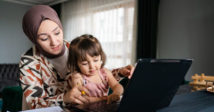 A woman in hijab sits in front of a computer. Her daughter, seated on her lap, pushes a button on the keyboard.