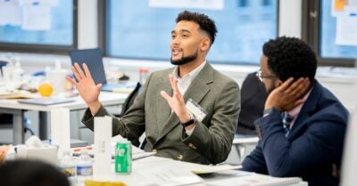 Man in a blazer gestures while speaking during a small group discussion in a modern office, as a colleague listens nearby with hand on chin.