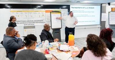 A facilitator stands at the front of a conference room, pointing to a flip chart while leading a training session on factor analysis. Participants seated around tables listen and take notes as a presentation is displayed on a large screen and workshop materials line the walls.